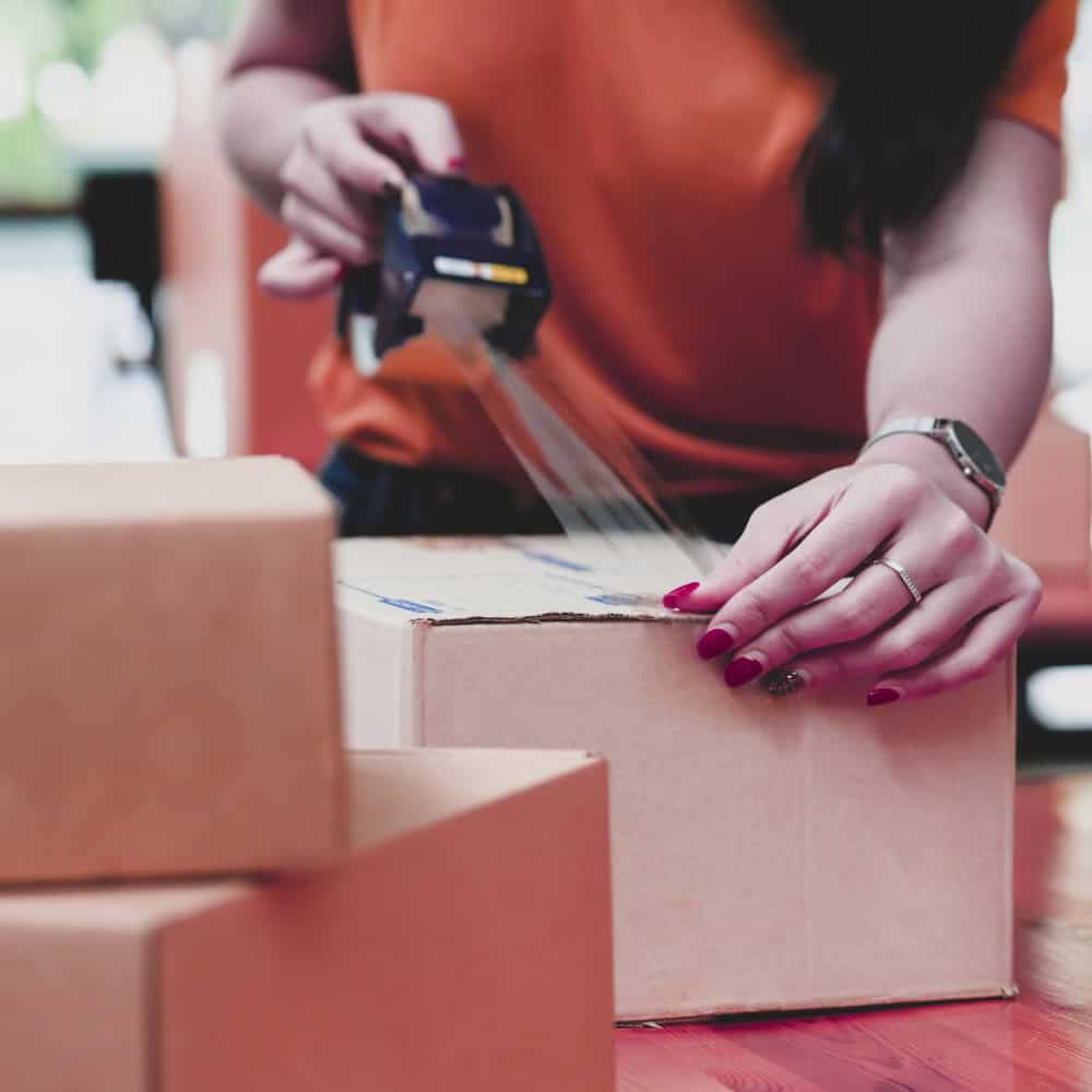 A person applying packing tape on a cardboard box, with additional boxes in the foreground.